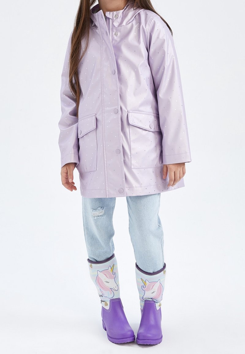 Lavendelfarbene, wasserdichte Jacke mit Kapuze, zwei vorderen Taschen und einem Punktmuster. Kombiniert mit hellblauen Jeans und Regenstiefeln mit Einhornmotiv.