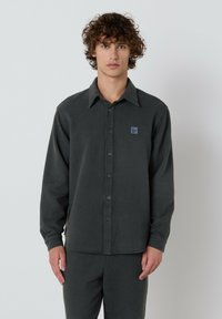 Mladenič s kodrastimi lasmi, oblečen v temno sivo srajco s gumbi in usklajenimi hlačami, stoji ob enobarvni svetli ozadju.