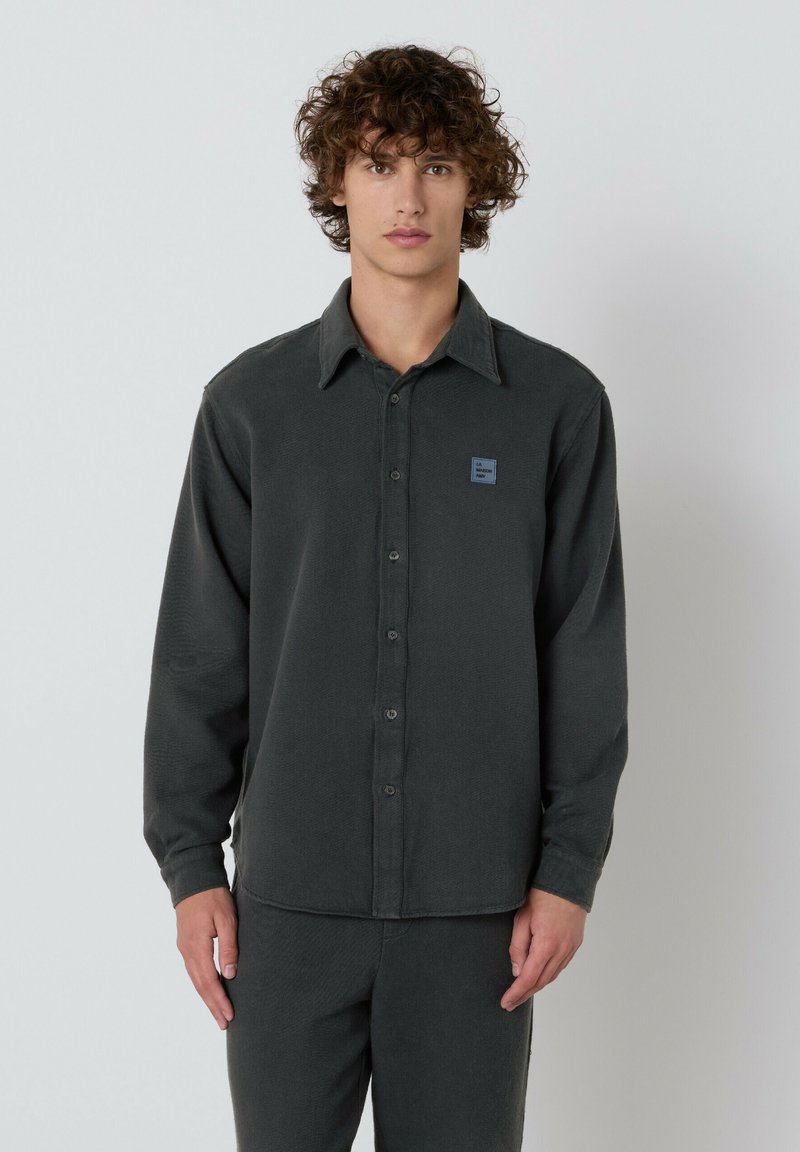 Mladenič s kodrastimi lasmi, oblečen v temno sivo srajco s gumbi in usklajenimi hlačami, stoji ob enobarvni svetli ozadju.