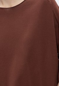 Gros plan d'une personne portant un pull tricoté marron uni avec un large col et des coutures visibles à l'épaule et à la manche.