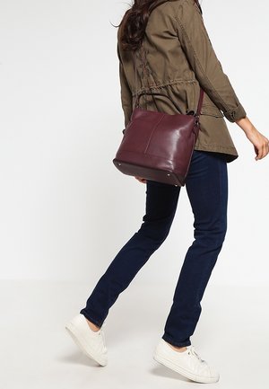 Donna che cammina con sneakers bianche, jeans blu scuro, giacca verde oliva, porta una borsa a tracolla in pelle rosso scuro.