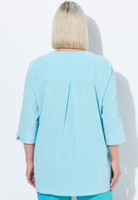 Lichtblauw shirt met een geplisséerde achterkant, driekwart mouwen en verticale strepen. Gemaakt van een geweven stof.