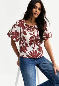 Blouse blanche avec un grand imprimé floral rouge, manches bouffantes, encolure froncée et détail à nouer. Associée à un jean bleu, assise sur un tabouret.