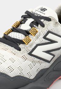 New Balance sneaker met een beige mesh bovenwerk, zwarte veters en gele en zwarte gemusterde veters accent. Dikke grijze zool met rode accenten.