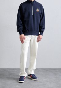 Man wearing navy half-zip sweatshirt with emblem, white jeans, and navy sneakers with red and white stripes, standing on gray floor.