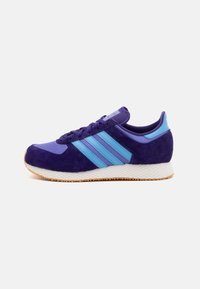 Violetiniai zomšiniai sportbačiai su mėlynomis dryžuotomis detalėmis, apvalia nosimi ir struktūrizuota balta guma. Su žemu profiliu ir paminkštintu apykakle.
