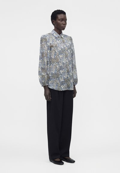 Femme portant un chemisier à manches longues bleu et beige à motif floral, associé à un pantalon large noir et des chaussures plates noires, debout sur un fond blanc.