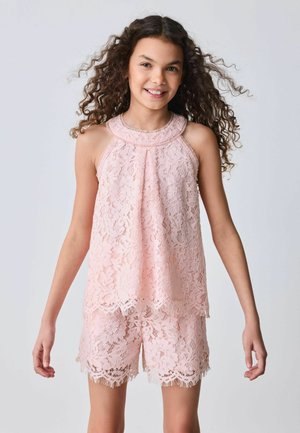 Tüdruk lokkis juustega, kandes varrukateta roosat pitsist topi ja sobivaid lühikesi pükse, naeratav, seisab kergelt sirutatud kätega.