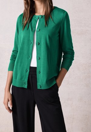 Kobieta ubrana w zielony kardigan na guziki, na biały top i czarne spodnie, z jedną ręką w kieszeni, na jasnym, neutralnym tle.