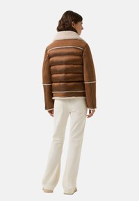 Braune Puffjacke mit einem Lammfellkragen, ausgestattet mit einem gesteppten Design, kontrastierenden weißen Nähten und einer boxy Passform. Tragen mit cremefarbenen Schlaghosen.