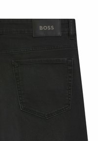 Jeans in denim nero con una texture liscia, dotati di una tasca a toppa e di un'etichetta rettangolare in rilievo con la scritta "BOSS" sulla vita.