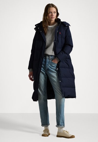 Cămașă lungă puffer de navy cu glugă, dotată cu nasturi capcană, design matlasat și un logo mic pe piept, asortată cu blugi bleu deschis și adidași.