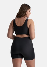 Kvinna med brunt hår iförd en svart ärmlös crop-top och högmidjade svarta cykelshorts, står med ryggen vänd mot kameran.