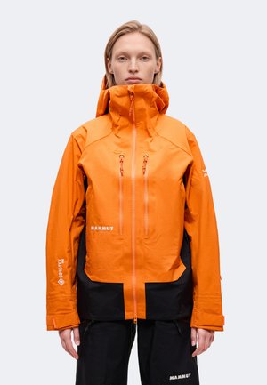 Un mannequin est de face, vêtu d’une veste imperméable orange Mammut à capuche haute et d’un pantalon noir, sur un fond clair uni.