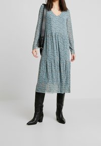 Długa, warstwowa sukienka w jasnoniebieskim materiale z czarnymi i brązowymi wzorami kwiatowymi, zestawiona z czarnymi kozakami do kolan i czarną torebką na ramię.