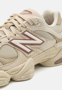 Beige athletic shoe with mesh and leather materials, featuring a textured sole, cream laces, and a maroon and white logo on the side.