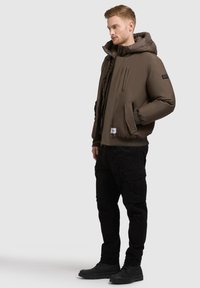 Veste bomber isolante marron avec capuche, dotée d'une fermeture éclair sur le devant, de poches latérales et de poignets côtelés, associée à un pantalon cargo noir et des bottes.