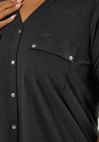 Zwarte blouse met V-hals, twee voorzakken en zilveren drukknopen. Zachte textuur met een relaxte pasvorm.