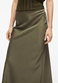 Μια σατέν φούστα ελιάς με λεία υφή και ριχτό σχέδιο. Η καμπυλωτή πλευρική ραφή προσφέρει διακριτική λεπτομέρεια.