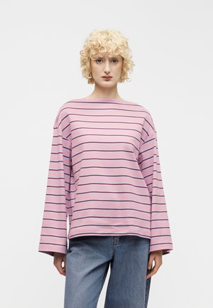 Mlajša oseba s kodrastimi svetlo rjavimi lasmi, oblečena v rožnato dolgo majico z ozkimi vodoravnimi črtami in modre kavbojke, stoji na belem ozadju.