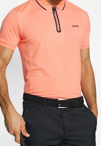 Man med korallfärgad zip-up polo-tröja med "BOSS"-logotyp, svart bälte och svarta byxor, håller en golfklubba med en hand i fickan.