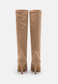 Une paire de bottes stiletto en suède beige, arrivant au genou, présentées de dos sur un fond blanc uni.