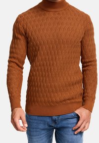 Homme portant un pull à col roulé texturé couleur rouille et un jean bleu, debout avec une main dans la poche, sur un fond blanc.