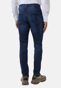 Tmavě modré džínové kalhoty slim fit, s černou koženou nášivkou na pásku a dvěma zadními kapsami. Nosené s béžovými botami.