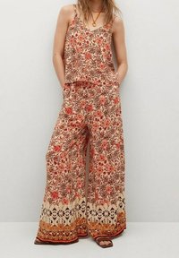 Combinaison à motifs floraux crème, ornée de designs rouges et marron, avec un pantalon à jambes larges, des bretelles spaghetti réglables et des poches sur les côtés.