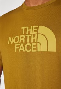 Dzeltenbrūnas īsām piedurknēm T-krekls ar "THE NORTH FACE" logo gaismas dzeltenā krāsā, novietots uz teksturētā auduma fona.