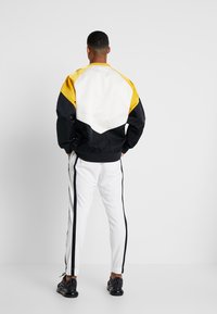 Flerfärgad jacka med gula, vita och svarta paneler; kombinerad med vita byxor med svarta sidostråk; mjukt material, elastiska muddar.