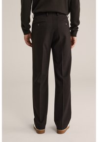 Mand iført sorte skræddersyede bukser og mørk sweater, stående med hænderne nær hofterne, set bagfra mod en neutral baggrund.