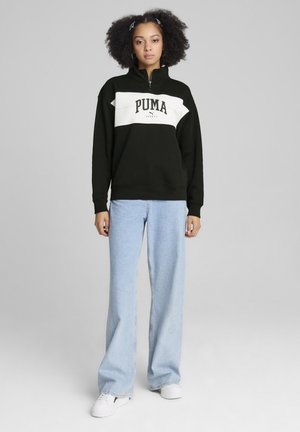 Schwarz-weiße Zip-Hoodie mit "PUMA"-Logo, kombiniert mit hellblauen weit geschnittenen Jeans und weißen Sneakers. Glatte Stofftextur.