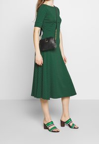 Kvinna som bär en grön och svart randig midiklänning, bär en svart crossbody-väska och har på sig gröna, svarta och vita klacksandaler.