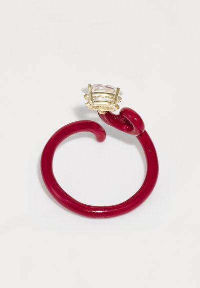 Roter Kunststoffring mit geschwungenem Design, gekrönt von einer goldenen Fassung, die einen klaren, facettierten, rechteckigen Stein hält. Glatte Oberfläche, glänzende Textur.