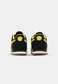 Une paire de baskets noires et jaunes vue de l'arrière sur une surface blanche unie.