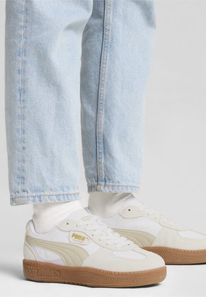 Białe i beżowe sneakersy Puma z gumowymi podeszwami, noszone z białymi skarpetkami i jasnoniebieskimi jeansami na jednolitym, jasnym tle.