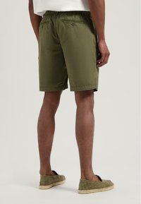 Olivgrüne Shorts mit elastischem Bund, zwei Gesäßtaschen mit Knöpfen und geradem Schnitt. Getragen mit olivfarbenen Slip-On-Schuhen.