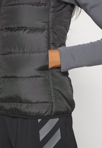 Svart pufferväst med sömmade paneler, sidoficka och grå långärmad topp. Har mörk tyg och slät yta.