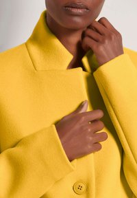 Geel wollen jas met een hoge kraag en lange mouwen, voorzien van een knoopdetail bij de manchetten en een enkele knoopsluiting.