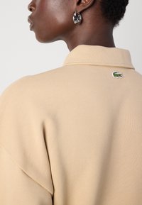 Polo shirt color beige con texture a coste e logo Lacoste ricamato in verde sul retro. Modello con orecchini a cerchio argentati.