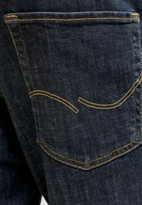 Približen posnetek zadnjega žepa temno modrih kavbojk iz jeansa z rumenim šivom v kontrastu in ukrivljenim dekorativnim detajlom.