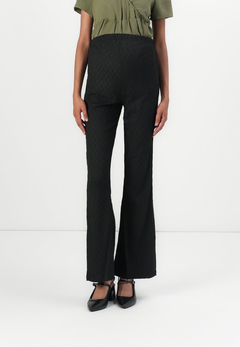 Pantalons noirs évasés avec un motif texturé et une taille haute, fabriqués en tissu tissé, associés à des chaussures en cuir verni noires.