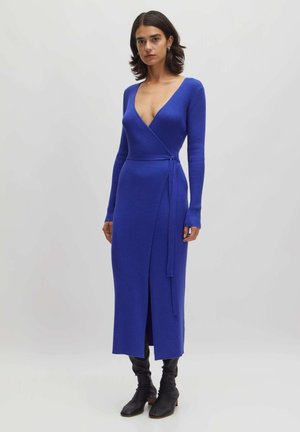 Langärmliges Wickelkleid mit tiefem V-Ausschnitt aus geripptem blauen Strick, mit seitlichem Bindeband und einem Oberschenkelspalt. Kombiniert mit knielangen schwarzen Stiefeln.