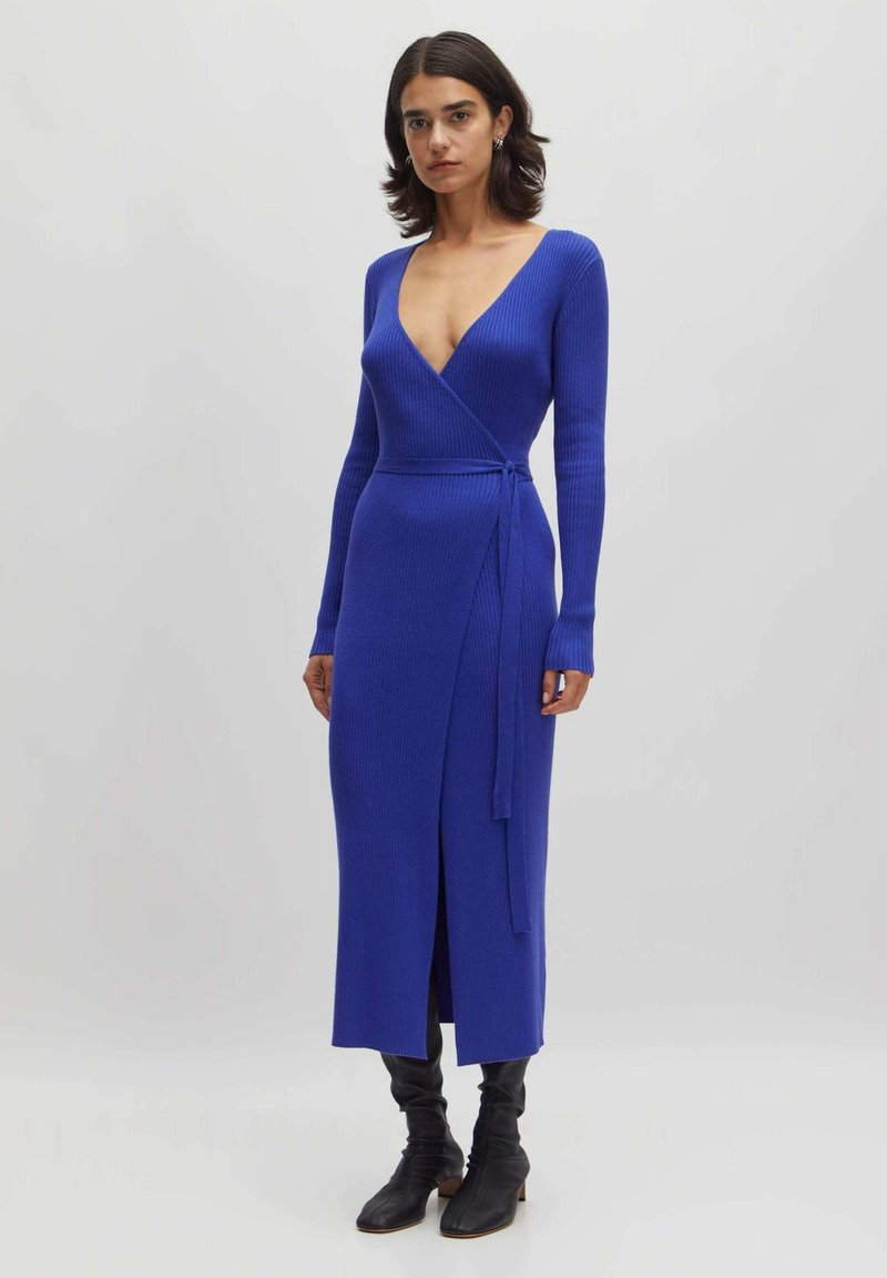 Langärmliges Wickelkleid mit tiefem V-Ausschnitt aus geripptem blauen Strick, mit seitlichem Bindeband und einem Oberschenkelspalt. Kombiniert mit knielangen schwarzen Stiefeln.