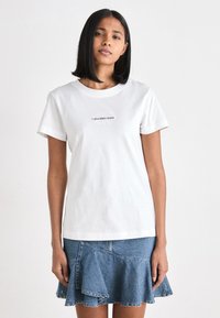 Maglietta in cotone bianco con maniche corte, caratterizzata da un logo nero "Calvin Klein Jeans" sul davanti. Indossata con una gonna di jeans con volant.