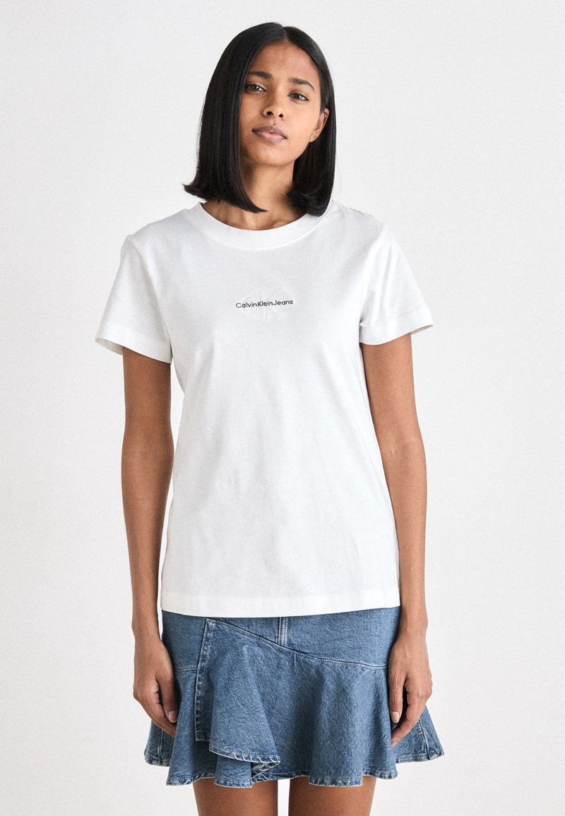 Maglietta in cotone bianco con maniche corte, caratterizzata da un logo nero "Calvin Klein Jeans" sul davanti. Indossata con una gonna di jeans con volant.
