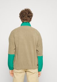 Chaqueta de forro polar beige con cuello y mangas de nailon verde. Presenta un ajuste relajado y puños elásticos. Textura suave, diseño minimalista.