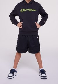 Černá mikina s kapucí a zeleným logem "Champion", doplněná černými síťovanými šortkami a černými teniskami s modrými detaily.