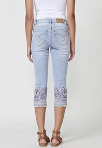Light blue cropped jeans with embroidered hem featuring floral patterns, frayed edges, and contrasting stitching, paired with brown sandals.